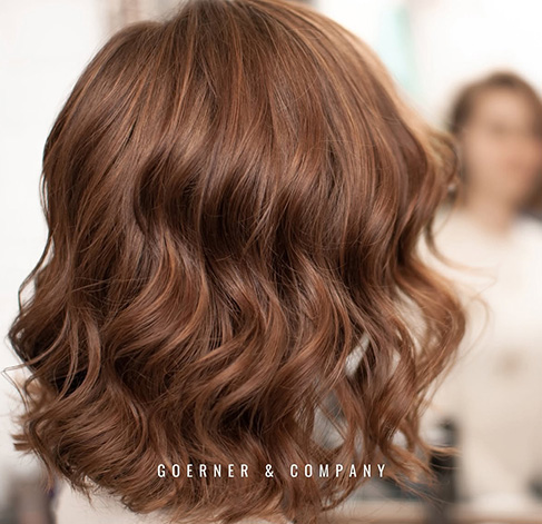 Ansicht eines Hinterkopfes. Die braunen Schulterlangen Haare fallen in weichen Wellen.