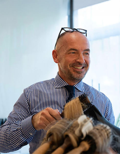 Friseur Goerner bei der Arbeit. Er schaut lächelnd aus dem Bild. Am unteren Rand ist der Kopf einer Person zu erkennen, dessen Haare er gerade föhnt.