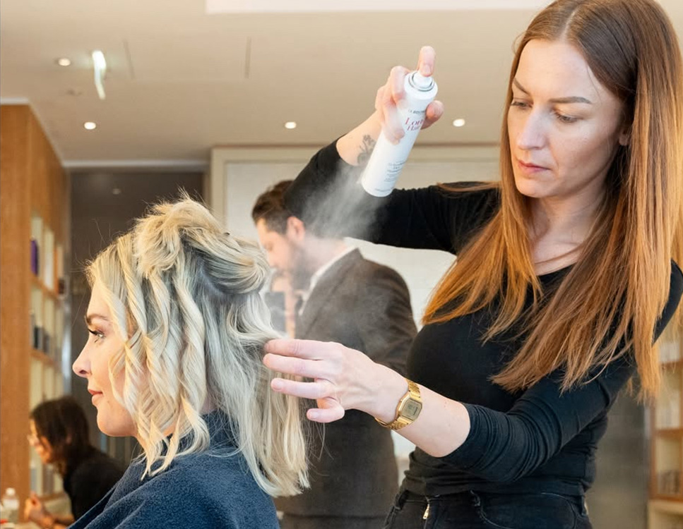 Eine Friseurin steht hinter einer Frau und stylt mit einem Spray die blonen lockigen Haare